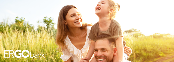 Smiling family outside in a field with mom next to father who is carrying their daughter on his shoulders