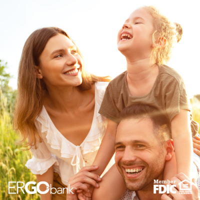 Smiling family outside in a field with mom next to father who is carrying their daughter on his shoulders