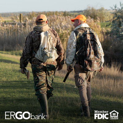 Two men in hunting gear walking outdoors with a dog